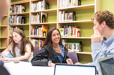 Two girls and a boy laugh and work in the Upper School library in front of a wall of books.