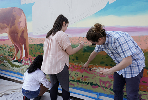 Artists work on outdoor mural of a howling coyote on a vintage postcard