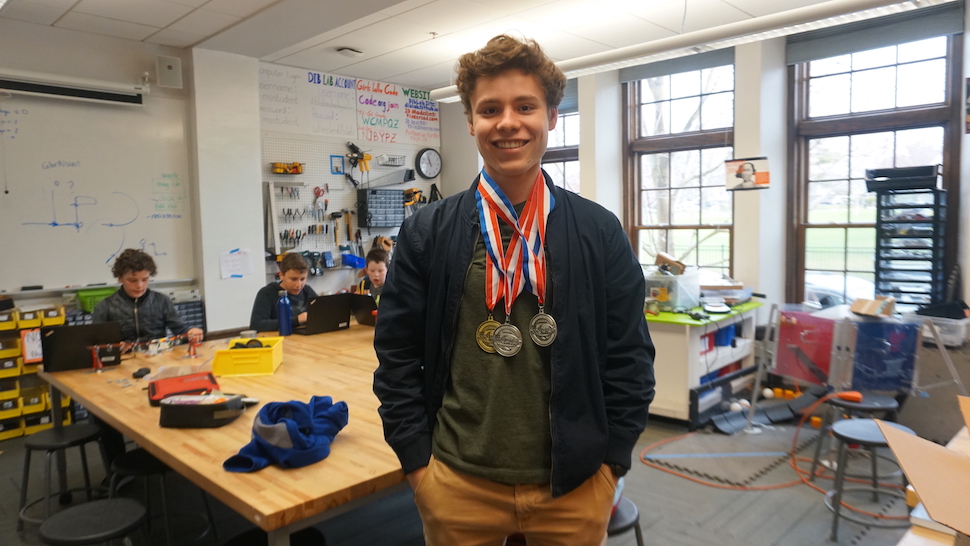 A high school student smiles at the camera while wearing three medals from a Science Olympiad.