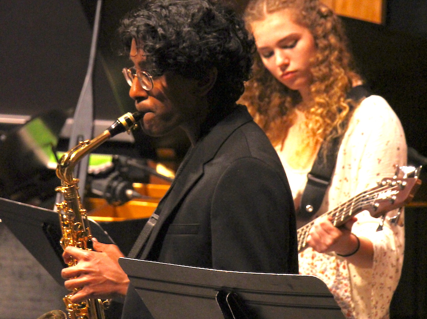 A boys plays saxophone and a girl behind him plays guitar at a performance.