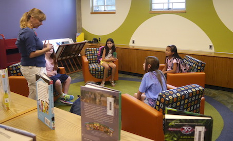 A librarian talks to four students sitting in comfy chairs in a brightly decorated reading room of the library.