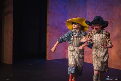 Two lower school girls wearing large hats and country-style dresses and aprons sing in a musical.