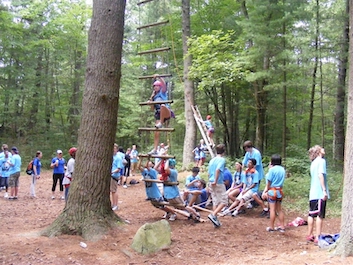 Large group of kids work on the various elements of a ropes course in the woods.