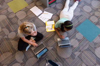 Two girls work on laptops on the floor of a classroom.