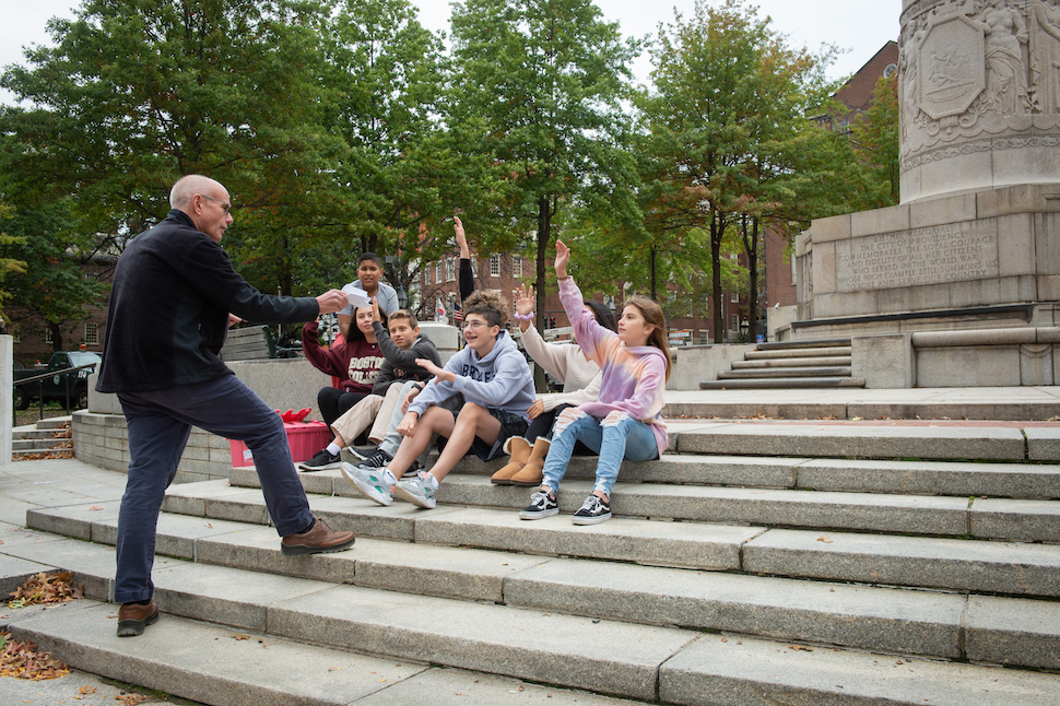 Joe Baer talks to a group of students, some of whom have their hands raised, as they sit on concrete steps near the water's edge in Providence.