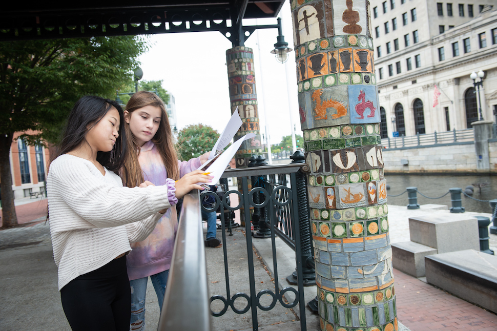 Two students look at their notes while standing near an art tile installation on a column near a bus stop in Providence.