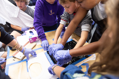 Students wearing purple gloves work with inky rollers in a maker's space.