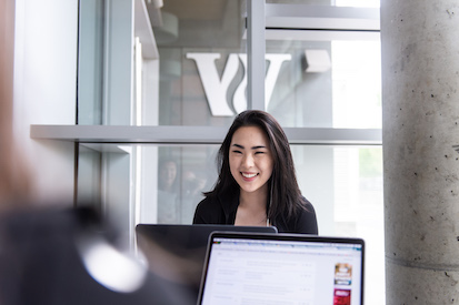 Smiling girl sits at laptop in front of window where you see a large silver W on the wall outside.