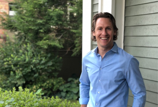Man with blue shirt standing on porch of older building.