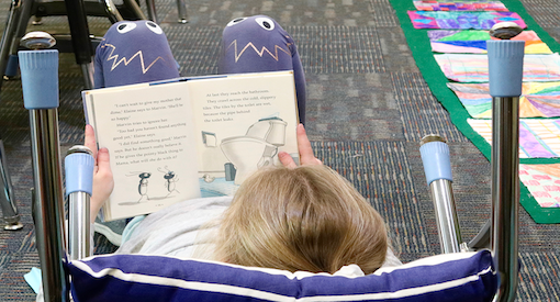 Student reclined in upside down desk chair reading a book on the floor.