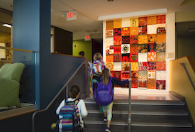 children with backpacks walk up stairs before a colorful tile installation.