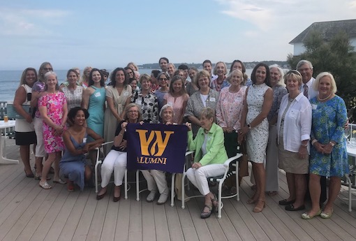 A group of men and women gather on a deck near the water holding a Wheeler flag.