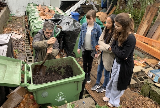 Three students watch a man turn over compost.