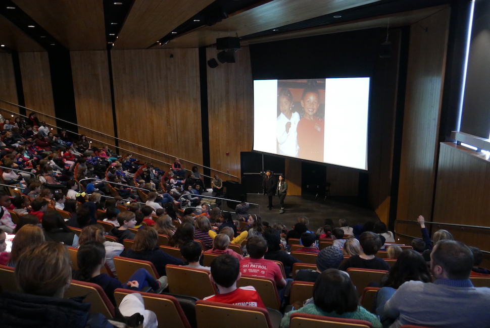 An auditorium of young students listen to two college students talk about their diversity and paths to the Ivy League while standing under a photo of themselves as children