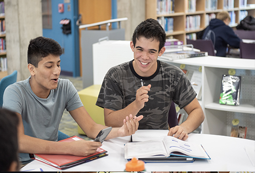 Two high school boys laugh while working in the library at a table.