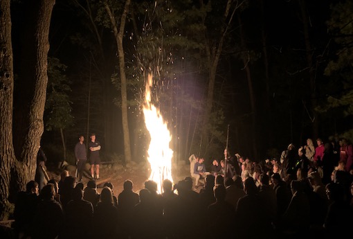 Students gather around a bonfire in the woods.