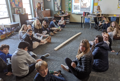A large group of 8th-grade and 3rd-grade students sit on a classroom floor surrounding a wall stud they will decorate together for Habitat for Humanity.