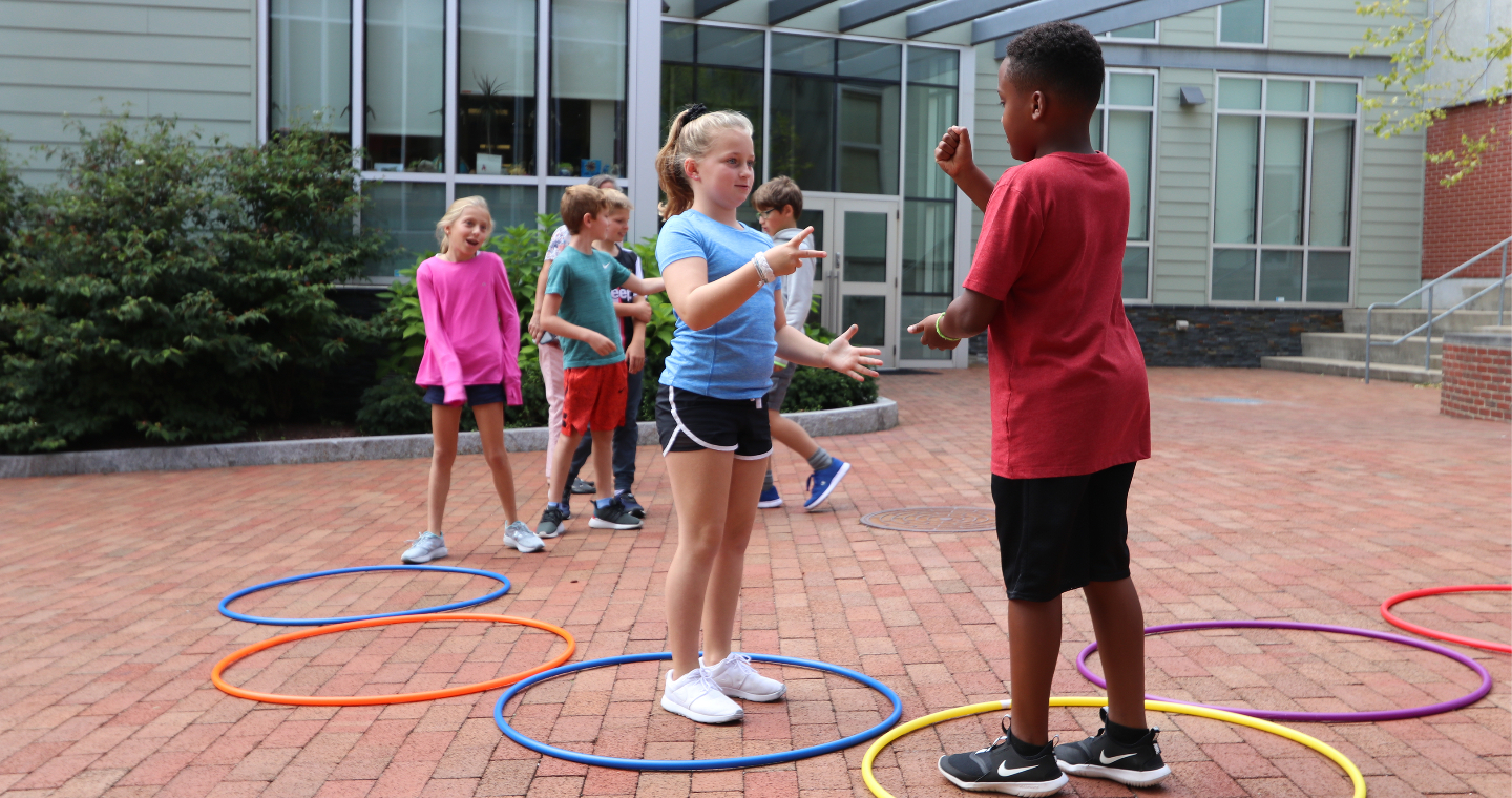 A group of children on a brick surface standing in hula hoops on the ground play Rock Paper Scissors.