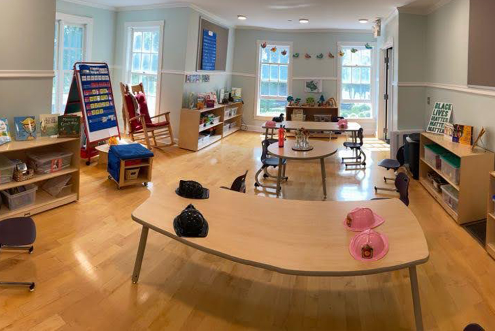 Long panoramic view of a new Nursery classroom with small tables and chairs, bright colorful flags, shelves, a rocking chair, and individual boxes of materials for each student.