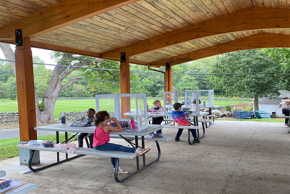 Students enjoy lunch under an outdoor pavilion with portable screens on each table for protection.