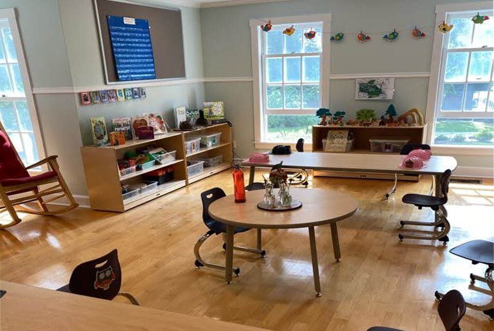 Sunny classroom view for Nursery students with small tables and chairs physically distanced, bright flags and books.