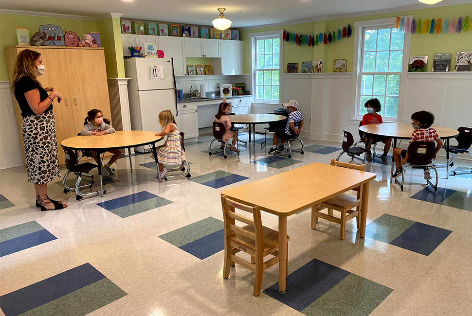 PreK students and their teacher wearing masks inside and distanced at tables in a colorful classroom at the Farm campus.