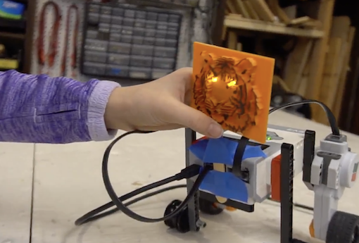 Little girl's hand reaches for 3D-printed tiger's face with illuminated eyes on a robot body.