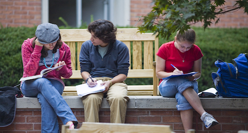 Three students sit on a bench doing homework.