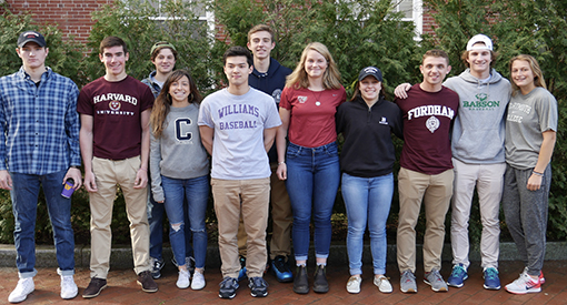 Eleven high school students wearing college sweatshirts standing in campus courtyard.