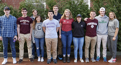 Eleven high school students wearing college sweatshirts standing in campus courtyard.