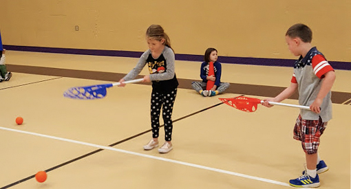 Young girl and young boy in Physical Education class practice using lacrosse sticks.