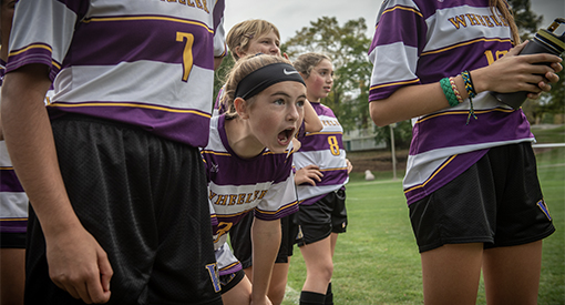 Group of Middle School girl athletes stand on the sidelines as one girl leans in to yell encouragement at the field.