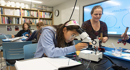 Middle School girl leans over a microscope while female teacher assists in Science lab.