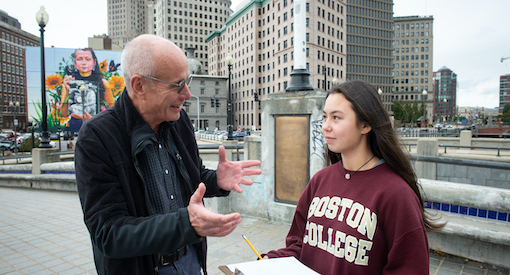 Male teacher speaks to 8th grade girl as they stand outside in Downtown Providence with large buildings and a colorful mural behind them.