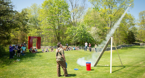 Teacher in overalls fires a classmade rocket into the air while groups of students stand back by a large red sculpture of an arch outside at The Wheeler Farm.