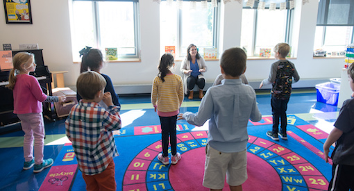 Music teacher works with group of young students standing on a colorful carpet in a music room.