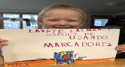 Little girl smiles broadly holding a sign she wrote in Spanish.