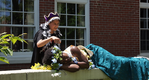 Boy dressed as a King leans over a girl pretending to sleep dressed as a princess in an outdoor enactment of a Shakespeare play.