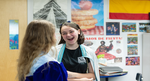 Two female teens standing laughing in a skit in Spanish Class. A bulletin board with a wall of posters in different languages is seen behind them.