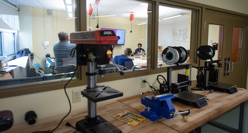 Machines sit in a maker space looking into a room with a class of teens and two teachers.