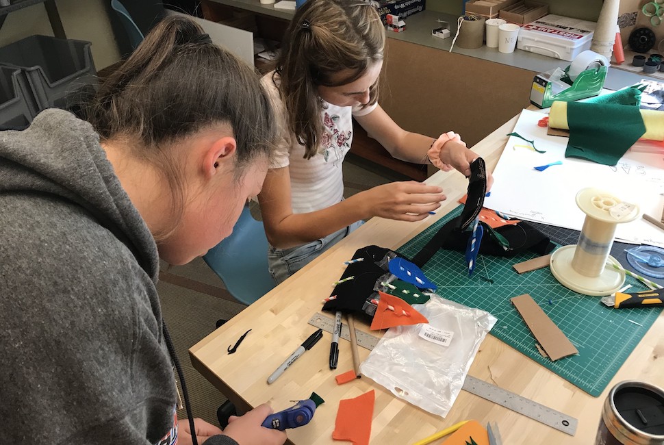 Two girls lean over tools in a design fabrication shop.