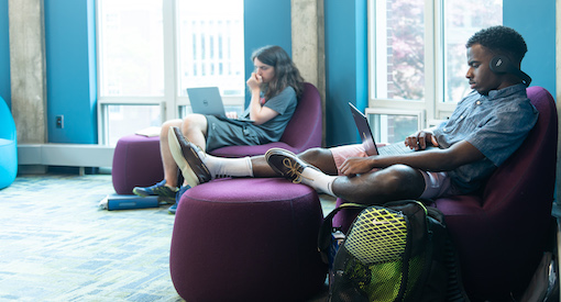 Two boys, one Black, one white read in comfy chairs in spacious library reading room.