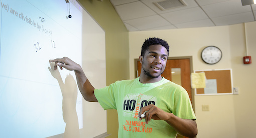 Black male teen smiles at the classroom while completely an equation on a board in math class.