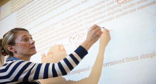 White female teacher writes a chemistry formula on a projected screen.