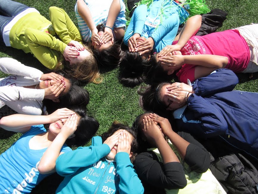 Group of young teens lie on ground in a circle with their hands over their eyes on a summer day.