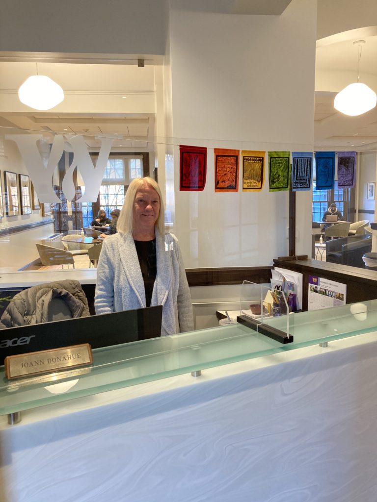 Woman stands smiling at reception desk where she has hung a set of peace flags. In the distance you see students studying at tables.