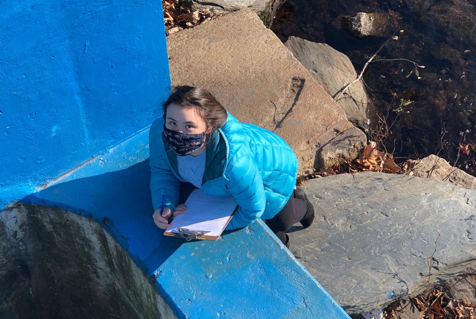 8th grade girl wearing mask looks up from her notetaking at a fish ladder in Providence.