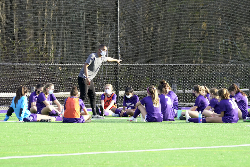 Girls soccer team sits on turf field while coach explains a play.