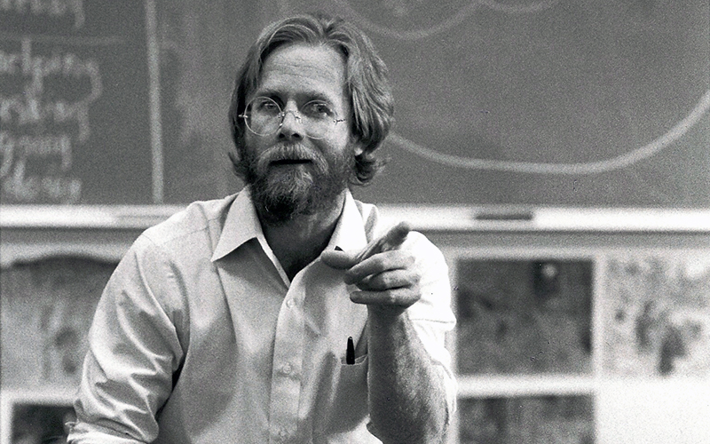 A black and white photo from 1987 of Mark Harris pointing and talking with someone off camera while sitting in a classroom with a blackboard behind him.