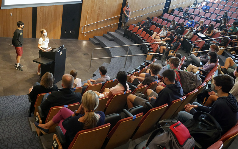 Ella Kulper speaks at the podium during Upper School assembly while Daniel Hu stands behind her. The backs of students, seated in auditorium chairs, are in the foreground.